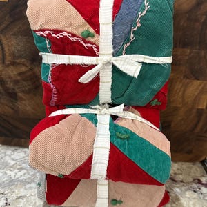 May include: A stack of colorful corduroy fabric pieces, sewn together in a patchwork design. The pieces are in shades of red, green, pink, and blue, and are tied together with white ribbon. The fabric has a quilted appearance with decorative stitching.