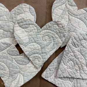 May include: A collection of heart-shaped quilted fabric pieces in shades of light blue and white. The fabric has a swirling, patterned design. The pieces are arranged on a brown paper surface, suggesting a craft or textile project.