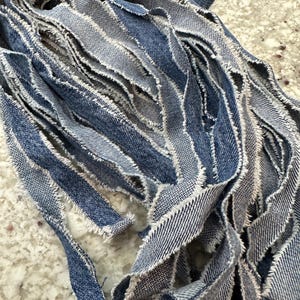 May include: Close-up of frayed denim strips in varying shades of blue. The strips have a raw, unfinished edge with visible white threads, creating a textured, layered effect. The fabric appears to be repurposed or upcycled.