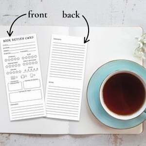 May include: Two white book review cards with black text sit on an open notebook. The front card has sections for title, author, and ratings. The back card has lines for thoughts and notes. A cup of tea on a blue saucer is in the image.