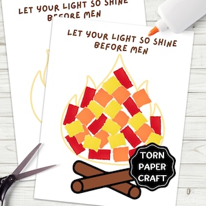 May include: Two white paper sheets with the text "LET YOUR LIGHT SO SHINE BEFORE MEN" and a torn paper craft campfire design. The campfire is made of red, orange, and yellow paper squares. Scissors and a glue bottle are also visible.
