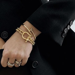 May include: A collection of gold jewelry, including a chain bracelet with a large clasp, a thin bangle, and a nail-shaped bracelet. Several rings adorn the fingers. The jewelry is set against a black blazer.