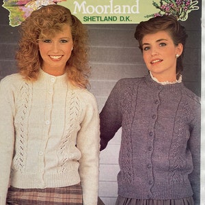 May include: Two women wearing knitted cardigans with a cable knit pattern. The cardigan on the left is white and the cardigan on the right is purple. The text "Patons Moorland Shetland D.K." is visible on the image.