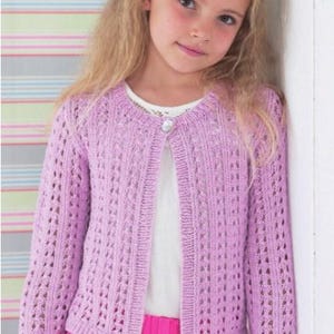 May include: A young girl wearing a light purple cardigan with a delicate openwork pattern. The cardigan has a button closure and is worn over a white shirt and a pink skirt.