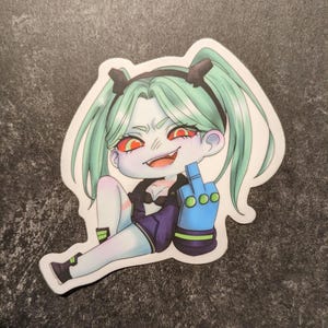 May include: A sticker featuring a cartoon character with green hair in pigtails, red eyes, and a mischievous grin. The character is giving the middle finger with a blue hand. The character is wearing a black top and purple shorts.