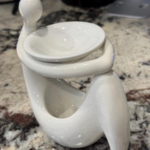 May include: White ceramic diffuser with a stylized human figure design. The diffuser has a round dish on top for wax melts or essential oils.