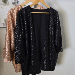 May include: Two sequined jackets hanging on wooden hangers. One is black, the other is a rose gold color. The jackets are open-fronted with long sleeves. The background is a light gray wall.