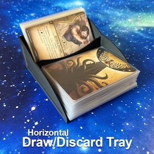 May include: A black plastic card holder with two compartments, designed to hold stacks of playing cards. The front compartment contains a deck with a tentacle illustration, while the back compartment holds a single card labeled Burning Daylight.