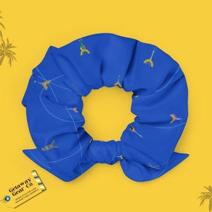 May include: Blue hair scrunchie with a repeating pattern of yellow palm trees on a white background. The scrunchie is tied in a knot and the text "Getaway Gear Co. Unique Styles for Your Unforgettable Adventures" is visible on a white and blue ticket.