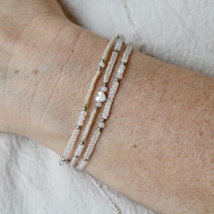 Könnte beinhalten: Drei cremefarbene Makramee-Armbänder mit kleinen, runden, silberfarbenen Perlen und einem kleinen, weißen herzförmigen Anhänger. Die Armbänder werden am Handgelenk getragen.