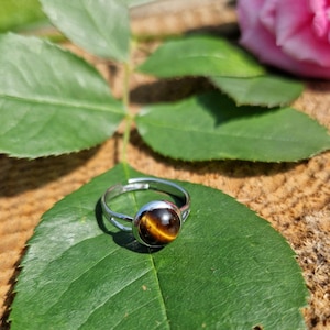 May include: A silver adjustable ring with a round, polished tiger's eye stone. The ring sits on a green leaf, with a blurred background of wood and a pink rose. The tiger's eye stone has brown and gold bands.
