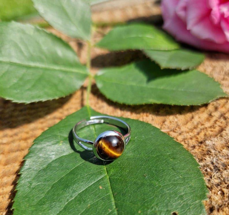 May include: A silver adjustable ring with a round, polished tiger's eye stone. The ring sits on a green leaf, with a blurred background of wood and a pink rose. The tiger's eye stone has brown and gold bands.