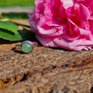 May include: A silver ring with a round, light green gemstone. The ring is set on a piece of wood next to a large, vibrant pink rose. The ring's design is simple, with a smooth, polished band.