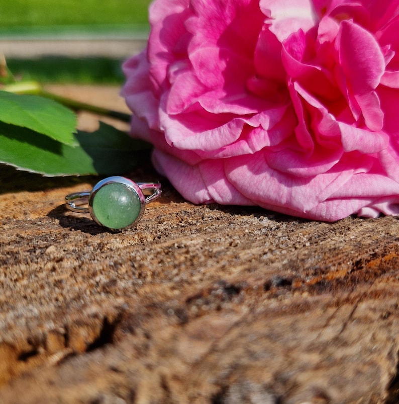 May include: A silver ring with a round, light green gemstone. The ring is set on a piece of wood next to a large, vibrant pink rose. The ring's design is simple, with a smooth, polished band.
