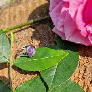 May include: A silver ring with a large, round, purple gemstone. The ring is set on a green leaf, with a pink rose and a wooden background. The ring is a piece of jewelry.