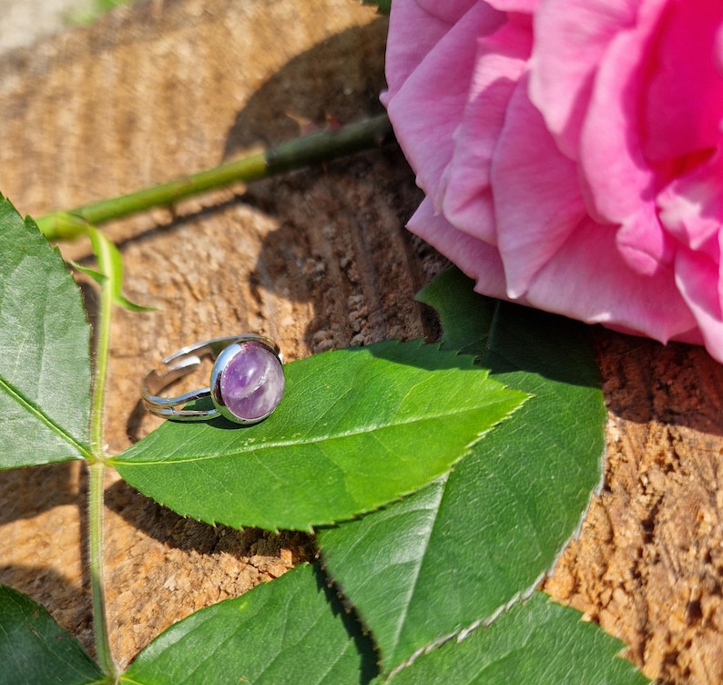 May include: A silver ring with a large, round, purple gemstone. The ring is set on a green leaf, with a pink rose and a wooden background. The ring is a piece of jewelry.