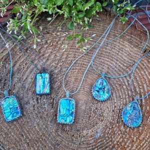 May include: Six necklaces featuring abalone shell pendants with iridescent colours. Each pendant has a unique shape, framed with a beaded border and strung on a matching cord. The necklaces are displayed on a textured wooden surface.