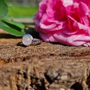 May include: A silver ring with a round, light pink gemstone. The ring is set on a textured, brown wooden surface, with a vibrant pink rose and green leaves in the background. The ring's design is simple and elegant.