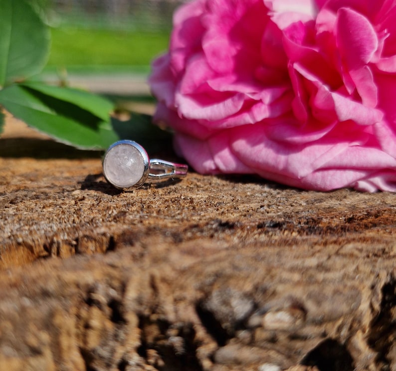 May include: A silver ring with a round, light pink gemstone. The ring is set on a textured, brown wooden surface, with a vibrant pink rose and green leaves in the background. The ring's design is simple and elegant.