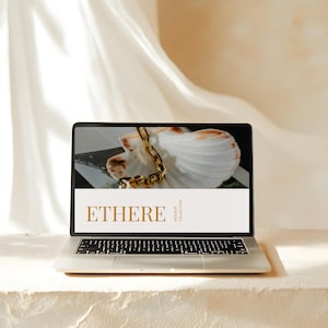 May include: A silver laptop displays a mockup with the word "ETHERE" in a gold font. The screen shows a seashell and gold chain. The laptop sits on a white surface, with a white fabric backdrop.