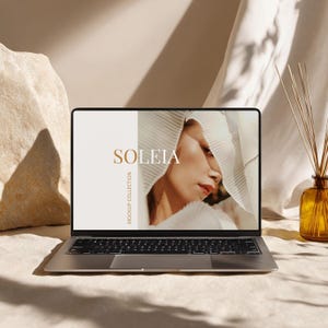 May include: A silver laptop displays a mockup with the word "SOLEIA" in gold lettering. The screen shows a woman wearing a white pleated fabric. The words "MOCKUP COLLECTION" are vertically aligned on the left side of the screen. A brown diffuser and a large rock are in the background.