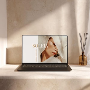 May include: A black laptop displays the word "SOLEIA" in gold lettering, with a woman's face on the screen. The laptop sits on a light-colored surface, with a diffuser and reeds in a brown glass bottle to the right.