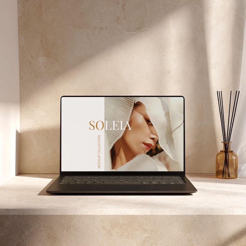 May include: A black laptop displays the word "SOLEIA" in gold lettering, with a woman's face on the screen. The laptop sits on a light-colored surface, with a diffuser and reeds in a brown glass bottle to the right.