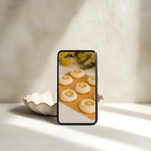 May include: A smartphone displays a close-up of several round cookies with a yellow filling and a dusting of white powder. The cookies sit on a brown surface. A decorative, off-white, scalloped bowl is in the foreground.