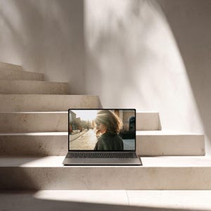 May include: A silver laptop with a black keyboard is open on a concrete staircase. The screen displays a person in a green jacket, with sunlight in the background. The steps are a light beige color, and the background is a white wall.