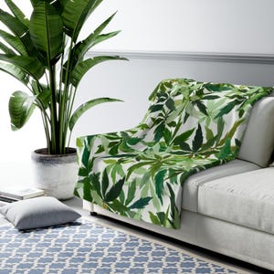 May include: A white sofa draped with a green leaf-patterned throw blanket. The blanket features a watercolor design of various green leaves. A large potted plant with green leaves is to the left of the sofa. A gray pillow and a blue patterned rug are also visible.