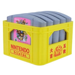 Peut inclure: Une caisse en plastique jaune remplie de cartouches de jeu Nintendo 64 grises. La caisse porte l'inscription "NINTENDO" en rouge et des caractères japonais. Les cartouches de jeu ont des étiquettes roses et colorées.