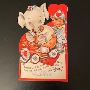 May include: Vintage Valentine's Day card featuring a cartoon elephant driving a red car, with a fireman in the back. The card has a heart-shaped design and the text "There's a fire in my heart, it's true, and the one who makes it burn is You!"