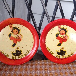 May include: Two red and white tin plates with a black and white illustration of a child jumping over a candle. The plates have the text "Jack Be Nimble" printed on them.