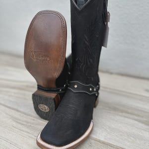 Puede incluir: Botas de vaquero de cuero negro con punta cuadrada y costuras decorativas. Las botas tienen un detalle de arnés con tachuelas plateadas y una suela de color claro. Se ve la suela de una bota, mostrando el nombre de la marca.