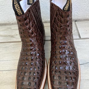 May include: A pair of brown leather cowboy boots with a square toe and a textured, crocodile-like pattern. The boots have a white and tan sole and the brand name "Carreta Boots" is printed on the pull tabs.