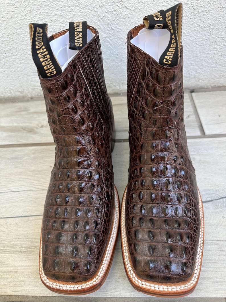 May include: A pair of brown leather cowboy boots with a square toe and a textured, crocodile-like pattern. The boots have a white and tan sole and the brand name "Carreta Boots" is printed on the pull tabs.