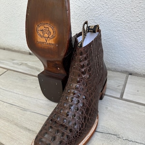 May include: Brown leather cowboy boots with a square toe and a textured, crocodile-like pattern. The soles are visible, with a logo and the words "Carreta Boots" stamped on them. The boots have a pull tab and a stitched design.