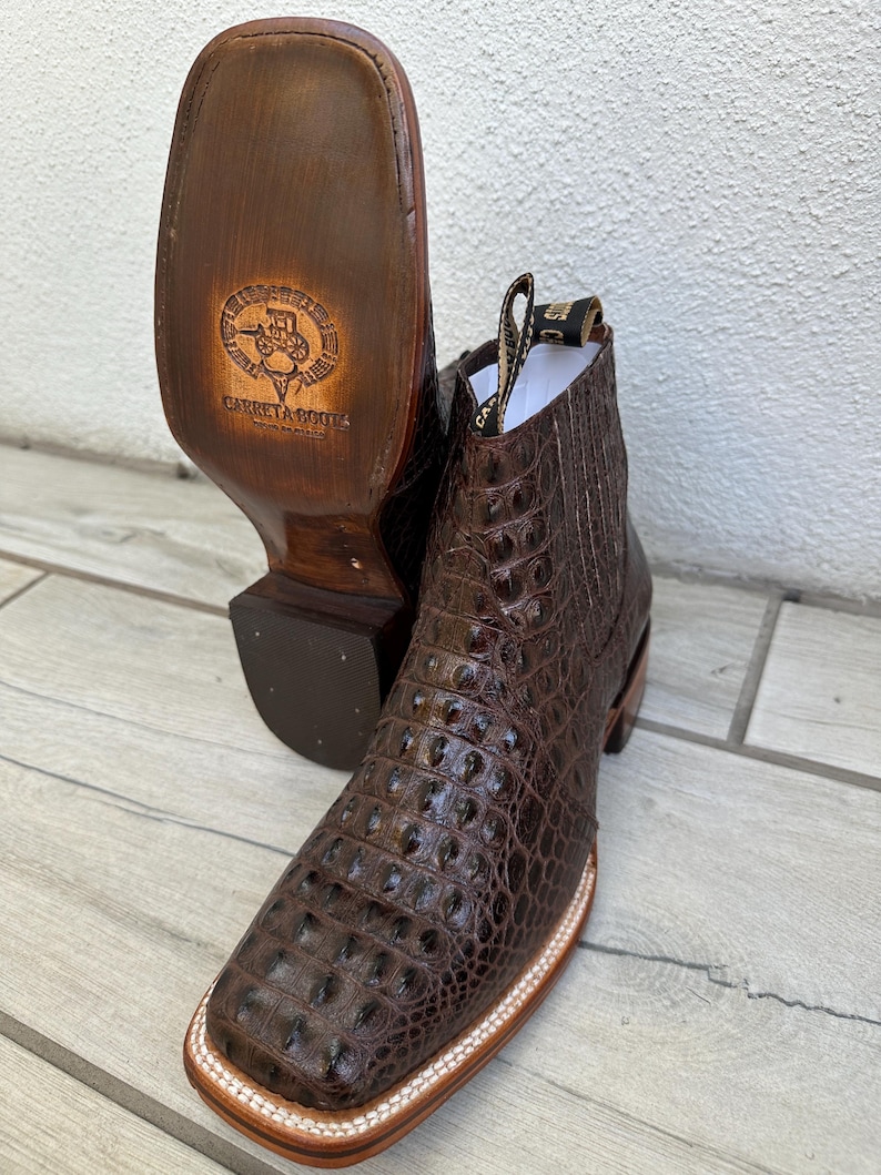 May include: Brown leather cowboy boots with a square toe and a textured, crocodile-like pattern. The soles are visible, with a logo and the words "Carreta Boots" stamped on them. The boots have a pull tab and a stitched design.