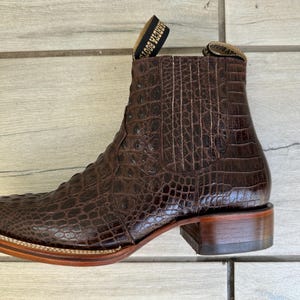 May include: Brown leather ankle boot with a crocodile skin texture. The boot has a square toe, a stacked heel, and a pull tab at the top. The sole is a light brown color with a stitched border.