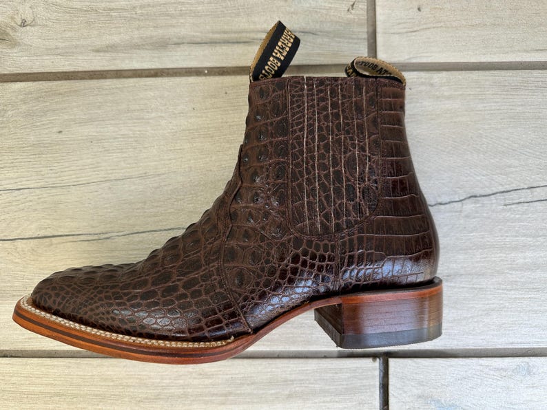 May include: Brown leather ankle boot with a crocodile skin texture. The boot has a square toe, a stacked heel, and a pull tab at the top. The sole is a light brown color with a stitched border.