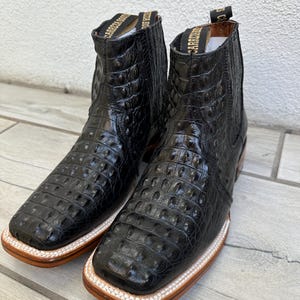 May include: A pair of black leather ankle boots with a square toe and a textured, crocodile-like pattern. The boots have a light brown sole and a white stitched border. The top of the boot has a gold label.