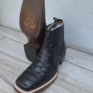 May include: Black leather cowboy boots with a square toe and a textured pattern. The boots have a brown sole and a stacked heel. The brand name "Carrera Boots" is visible on the pull tab. The boots are displayed on a light wood surface.