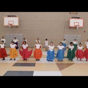 May include: A group of children in a gymnasium, wearing colorful traditional dance costumes. The costumes include white tops and vibrant skirts in red, orange, yellow, green, and blue. The children are standing in a line.