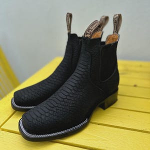 May include: A pair of black leather boots with a textured, snake-like pattern. The boots have a square toe, a low heel, and a white stitched border. The pull tabs at the top are tan with the brand name.