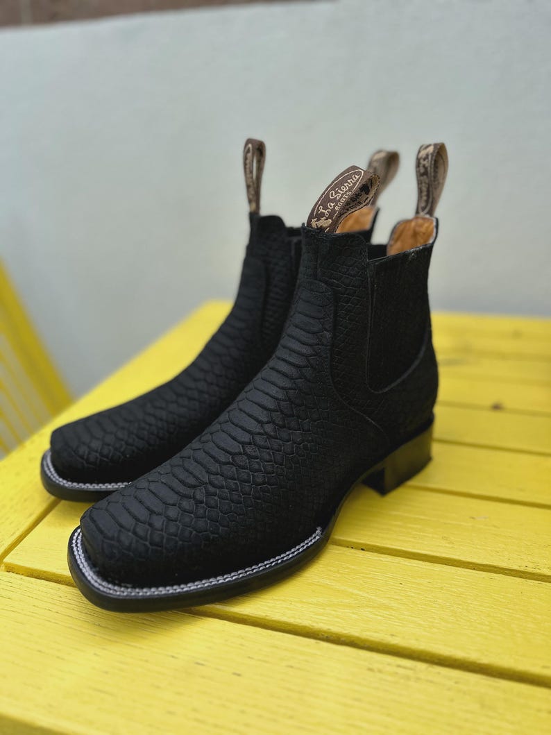 May include: A pair of black leather boots with a textured, snake-like pattern. The boots have a square toe, a low heel, and a white stitched border. The pull tabs at the top are tan with the brand name.
