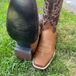 May include: A pair of brown leather cowboy boots with decorative stitching and a square toe. The boots have a dark brown upper and a lighter brown foot. The sole of one boot is visible, with the words "El General" and an eagle logo.