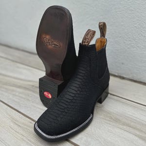 May include: Black leather cowboy boots with a square toe and a textured pattern. The boots have a pull tab and a white stitched border around the sole. The sole is brown and the heel is black.