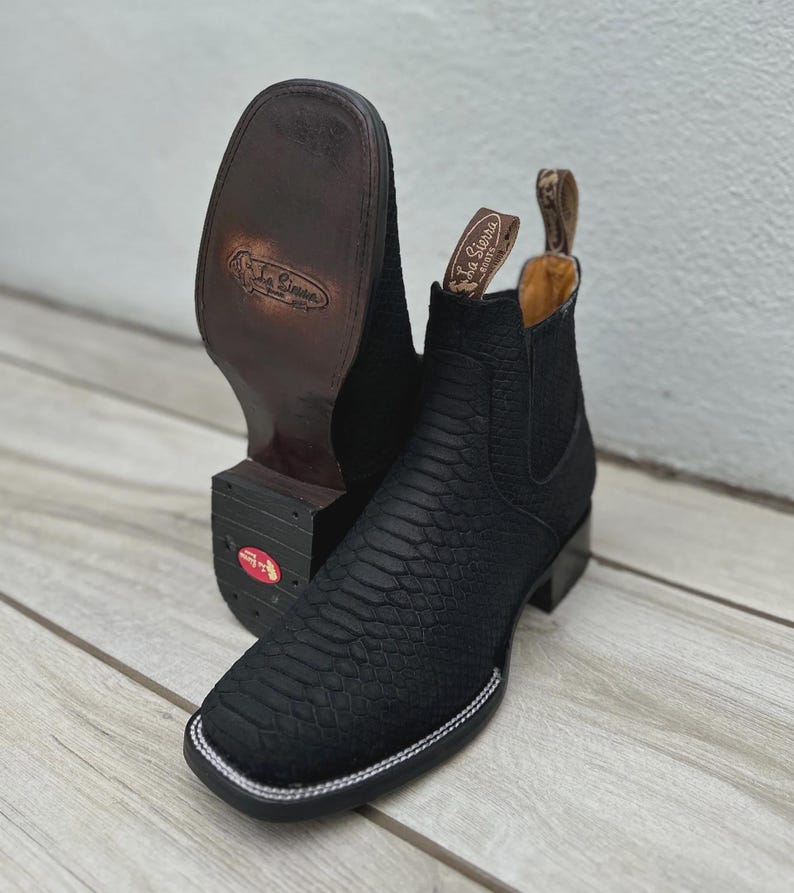 May include: Black leather cowboy boots with a square toe and a textured pattern. The boots have a pull tab and a white stitched border around the sole. The sole is brown and the heel is black.