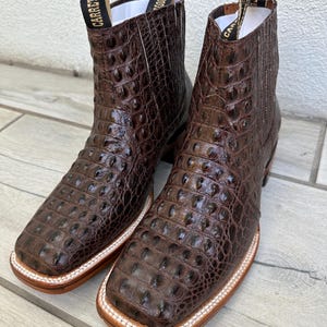 May include: A pair of brown leather ankle boots with a crocodile skin texture. The boots have square toes, pull tabs with text, and a white and tan sole. The boots are displayed on a light wood surface.