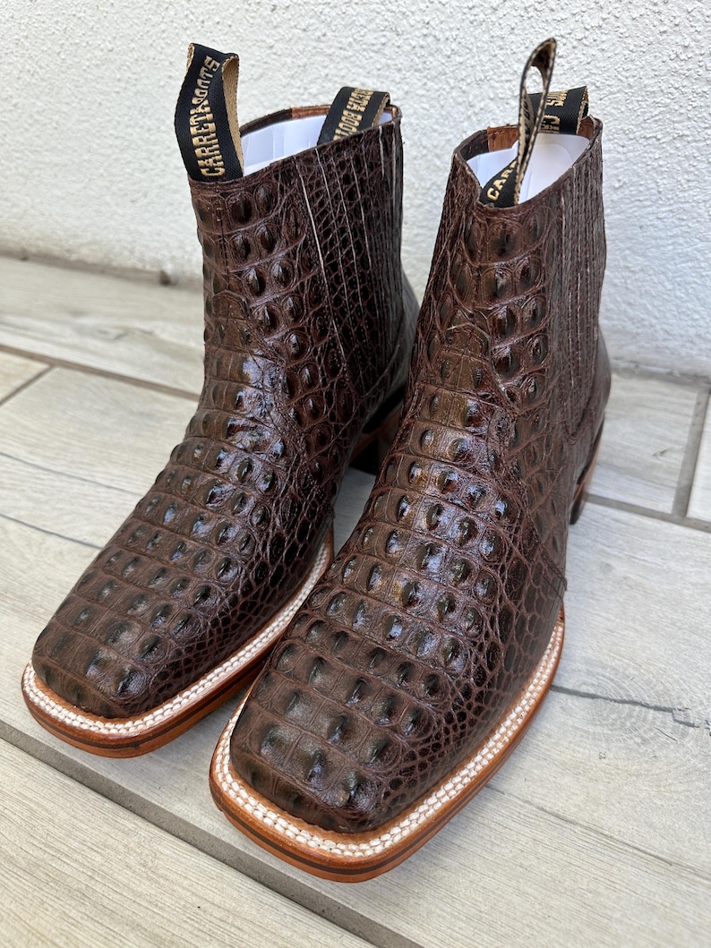 May include: A pair of brown leather ankle boots with a crocodile skin texture. The boots have square toes, pull tabs with text, and a white and tan sole. The boots are displayed on a light wood surface.